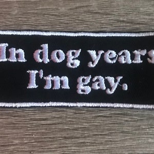 May include: Black embroidered patch with white stitching. The text on the patch reads "In dog years I'm gay."