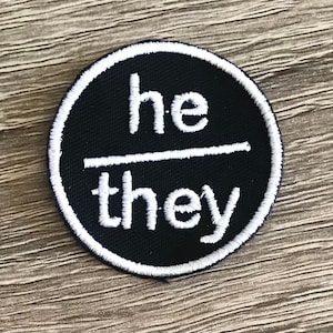 May include: Black and white embroidered patch with the words "he/they" in a circle.