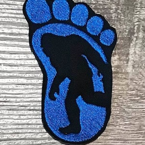 May include: Blue and black embroidered patch of a Bigfoot footprint with a silhouette of Bigfoot inside the footprint.