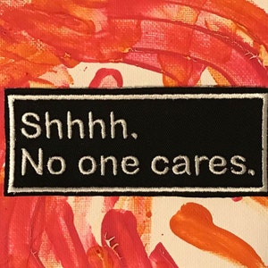 May include: A black patch with white stitching and the text "Shhhh. No one cares." on a red and orange painted background.