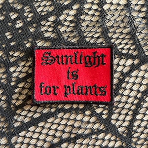 May include: A rectangular red embroidered patch with a black border. The patch features black text in a gothic-style font that reads Sunlight is for plants. The embroidery has a visible texture and is centered on the fabric surface.