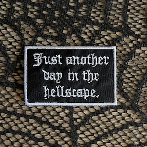 May include: A rectangular black embroidered patch with a white border. The patch features the white text "Just another day in the hellscape." written in a gothic-style font. The embroidery has a visible diagonal texture across the black surface.