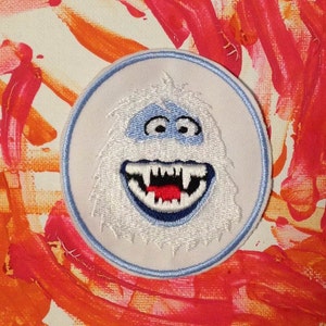 May include: White embroidered patch of a smiling snowman with a blue face and red mouth. The patch is circular and has a light blue border.