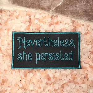 May include: Black embroidered patch with teal stitching. The patch reads "Nevertheless, she persisted."