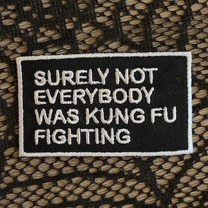 May include: A rectangular black embroidered patch with a white border and white stitched text that reads: SURELY NOT EVERYBODY WAS KUNG FU FIGHTING. The patch features a clean, bold font and a durable fabric construction with a finished edge.