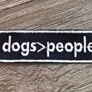 May include: Black and white embroidered patch with the text "dogs>people" on a black background.
