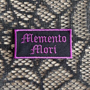May include: Black fabric patch with purple stitching. The patch says "Memento Mori" in a gothic font.