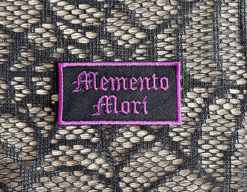 May include: Black fabric patch with purple stitching. The patch says "Memento Mori" in a gothic font.