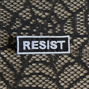 May include: A rectangular embroidered patch featuring a black background with the word RESIST stitched in bold white capital letters, surrounded by a thin white border, placed against a black lace fabric with a repeating floral pattern.