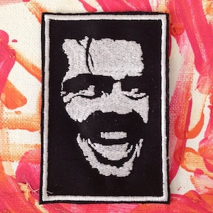 May include: A black and white embroidered patch of a smiling face with a mustache. The patch is surrounded by a white border.