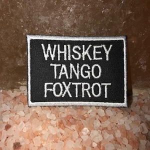 May include: A black rectangular patch with white embroidered text that reads "WHISKEY TANGO FOXTROT." The patch has a white border. The background is a light brown surface with pink and white crystals.