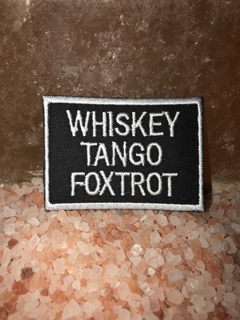May include: A black rectangular patch with white embroidered text that reads "WHISKEY TANGO FOXTROT." The patch has a white border. The background is a light brown surface with pink and white crystals.