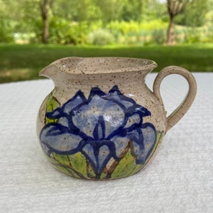 May include: A small, cream-colored ceramic creamer with a blue and green floral design. The creamer has a rounded body and a single handle.