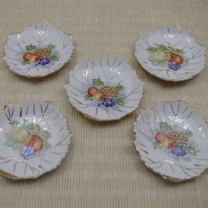 May include: Set of five white ceramic leaf-shaped dishes with gold trim. Each dish features a painted design of fruit, including a pineapple, grapes, and apples.