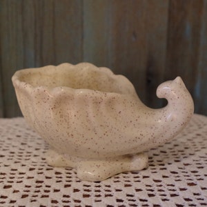 May include: A white ceramic planter shaped like a cornucopia with brown speckles throughout. The planter has a wide opening and a curved handle.