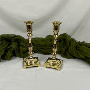 May include: A pair of gold metal candle holders with a square base and a fluted design. The candle holders are on a green fabric background.