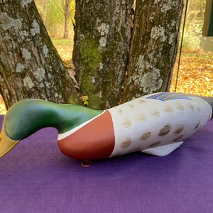 Puede incluir: Señuelo de pato de madera tallado a mano con pintura verde, marrón, blanco y azul. El pato está acostado sobre una superficie morada.