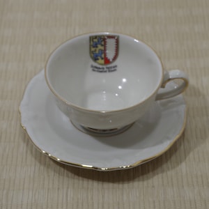May include: White porcelain teacup and saucer with a gold rim. The teacup has a coat of arms with the text "Schwäbisch Gmünd,  Architektur Stadt".