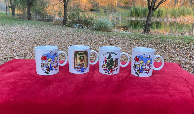 May include: Set of four white ceramic mugs with Christmas scenes. Each mug features a different scene with a house, a Christmas tree, and children. The mugs have blue, red, green, and yellow accents.