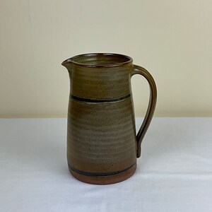 May include: A brown ceramic pitcher with a wide mouth and a handle. The pitcher has a brown glaze with a darker brown stripe around the middle.