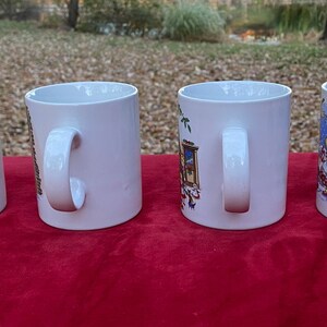 May include: Four white ceramic mugs with different holiday-themed designs. The mugs have white handles and are arranged in a row on a red surface.