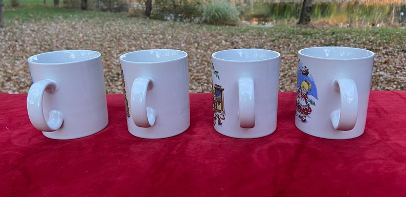 May include: Four white ceramic mugs with different holiday-themed designs. The mugs have white handles and are arranged in a row on a red surface.