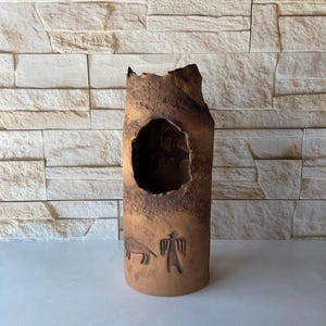 May include: A tall, brown ceramic vase with a large, irregular opening and carved primitive figures. The vase has a rough, textured surface and a cylindrical shape. The background is a light-colored stone wall.