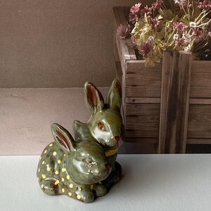 May include: A pair of ceramic bunny figurines in a sage green hue. One bunny is adorned with yellow polka dots. The bunnies are nestled together, with red accents on their ears and noses. A decorative item for Easter or spring.