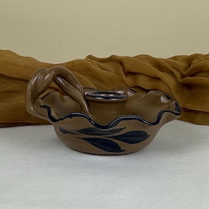 May include: A brown ceramic basket with a handle and a wavy rim. The basket has a dark blue leaf design on the side and a dark blue rim. The handle is a twisted design. The basket is on a white surface.