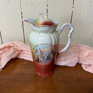May include: A vintage porcelain creamer or coffee pot with a lid. The pot is decorated with a colorful scene of people in a landscape. The pot is white with a red and green ombre glaze.