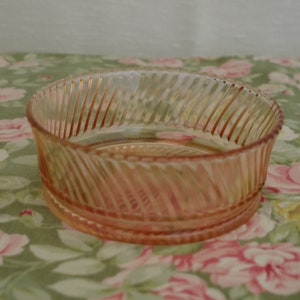 May include: A pink glass bowl with a swirl design. The bowl is empty and sitting on a floral tablecloth.