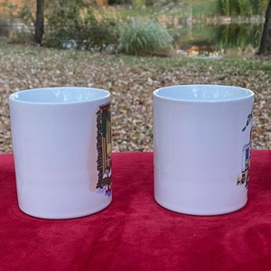 May include: Four white ceramic mugs with colorful illustrations of houses on them. The mugs are lined up in a row on a red surface.