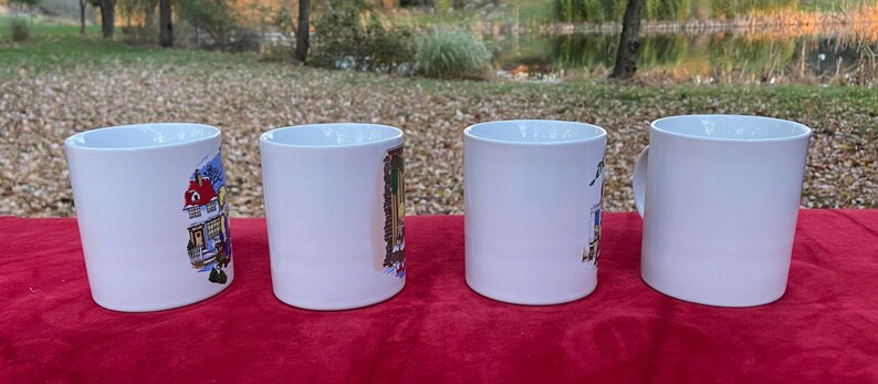 May include: Four white ceramic mugs with colorful illustrations of houses on them. The mugs are lined up in a row on a red surface.