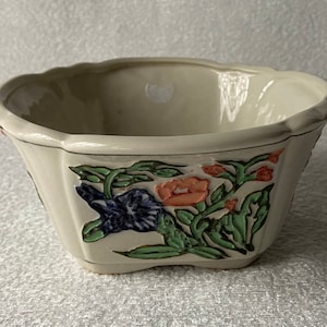 May include: A white ceramic planter with a scalloped rim and floral design. The planter features green leaves, pink and orange flowers, and blue flowers.