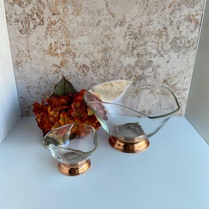 May include: Two clear glass bowls with a unique, asymmetrical shape. Each bowl sits on a copper-colored base. The bowls are displayed on a white surface with a patterned background and a bouquet of orange flowers.