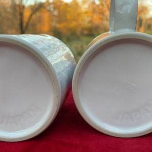 May include: Two white ceramic mugs with the word "JAPAN" printed on the bottom. The mugs are on a red surface.