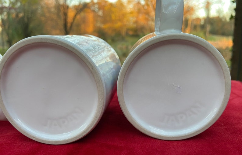 May include: Two white ceramic mugs with the word "JAPAN" printed on the bottom. The mugs are on a red surface.