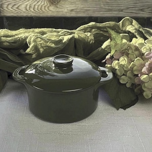 May include: A dark green ceramic cooking pot with a matching lid. The pot has two small handles and a round knob on the lid. The pot is sitting on a white surface with green fabric and flowers in the background.