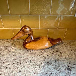 Puede incluir: Un señuelo de pato de madera tallado en una sola pieza de madera. El pato es marrón y tiene un acabado de madera natural. Es un señuelo de estilo tradicional con una forma realista.
