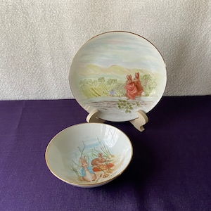 May include: Two white ceramic plates with a gold rim. One plate features a colorful illustration of two rabbits in a field. The other plate features a colorful illustration of two rabbits in a field with a blue sky.