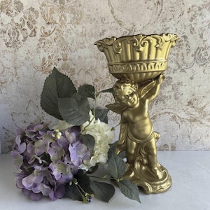 May include: A gold-colored cherub figurine holding a decorative bowl. The cherub is standing on a base, and the bowl has an ornate design. A bouquet of purple and white hydrangeas with green leaves is next to the figurine.