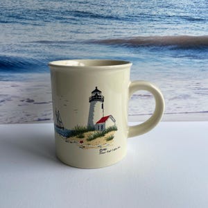 May include: Cream-colored ceramic mug featuring a coastal scene. The design includes a lighthouse with a red roof, a small white building, and a sailboat. The mug has a curved handle and a slightly flared rim. The background shows a blue ocean.