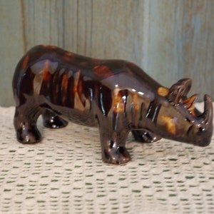 May include: A brown ceramic rhinoceros figurine with a glossy finish. The figurine is standing on a white crocheted surface.