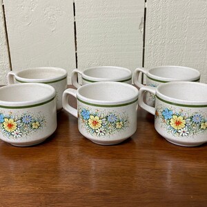May include: Six ceramic coffee cups with handles. The cups are off-white with a speckled texture and a green band around the rim. Floral designs in yellow, blue, and white decorate the front of each cup.