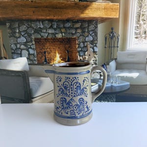 May include: A beige ceramic pitcher featuring a dark brown interior and a handle on the side. The exterior is decorated with blue hand-painted floral and heart-shaped patterns, including a small blue squiggle design near the base of the vessel.