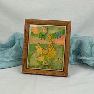 May include: A framed watercolor painting of a giraffe, a cat, a lion, and two hippos celebrating a birthday with a cake and balloons. The animals are wearing party hats and the background is a green field with a blue sky.