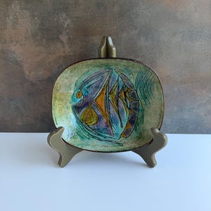 May include: An oval ceramic dish featuring a stylized fish illustration in shades of teal, purple, and yellow against a mottled green background. The dish has a dark brown rim and is displayed on a dark green plastic stand with three prongs.