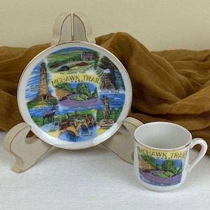 May include: A white ceramic saucer and teacup with a gold trim. The saucer features a colorful illustration of the Mohawk Trail in Massachusetts, with landmarks like the hairpin turn, the bridge of flowers, and Mt. Greylock Memorial. The teacup has a similar design, but only shows the hairpin turn.