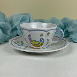 May include: A white teacup and saucer with a blue and yellow bird design. The bird is perched on a vine with green leaves and purple flowers.
