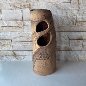 May include: A tall, cylindrical, tan-colored vase with two oval cutouts revealing a glimpse of a face. The vase has a textured surface with gold accents and geometric patterns. The vase is likely ceramic.